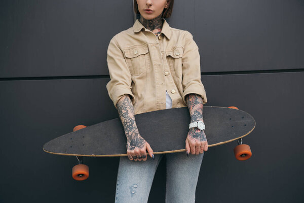cropped image of tattooed woman holding skateboard against black wall 