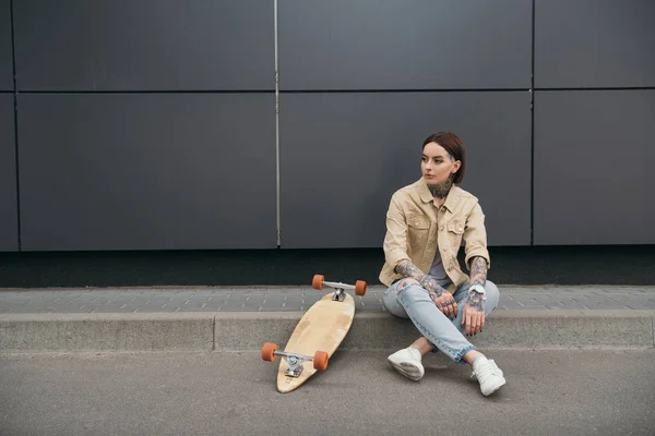 tattooed stylish woman sitting near skateboard at street