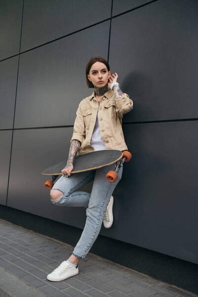 low angle view of tattooed woman holding skateboard against black wall 