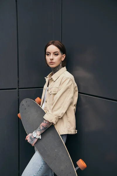 young tattooed woman holding skateboard against black wall