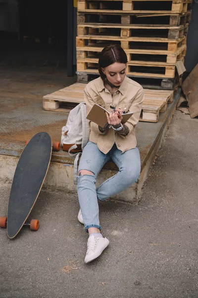 stylish tattooed woman with skateboard writing in textbook