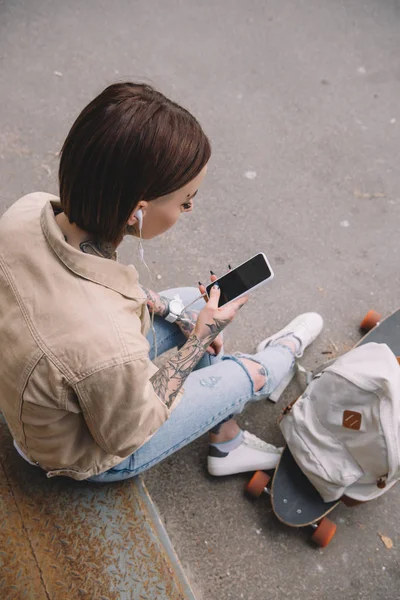 高角度视角的纹身妇女在耳机听音乐与智能手机附近的滑板和背包 — 免费的图库照片