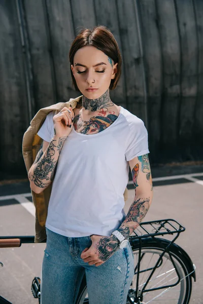 young tattooed woman with closed eyes holding jacket over shoulder near bicycle
