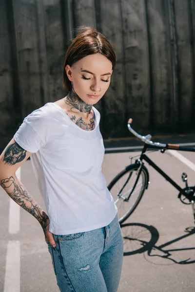 young tattooed woman with closed eyes posing near bicycle at street