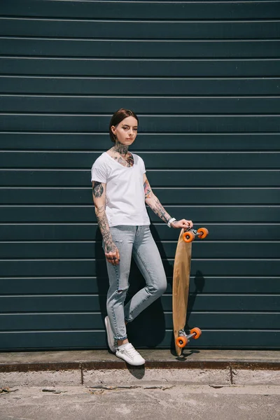 young tattooed woman standing with skateboard against black wall