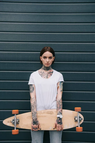 serious tattooed girl holding skateboard and looking at camera against black wall