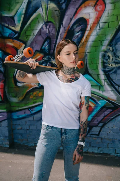 young woman with tattoos holding skateboard over shoulder near wall with colorful graffiti