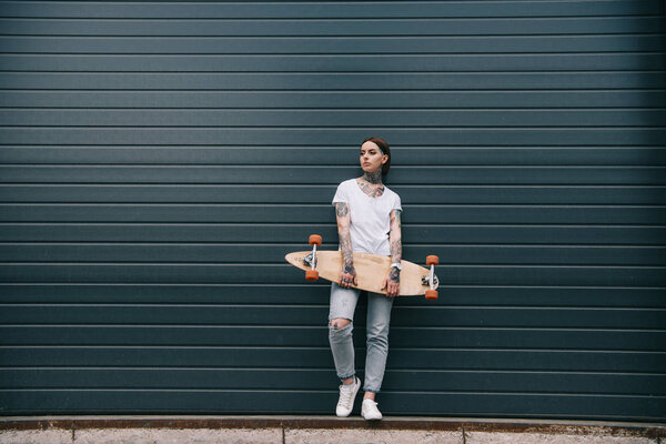 distant view of young woman with tattoos holding skateboard against black wall 