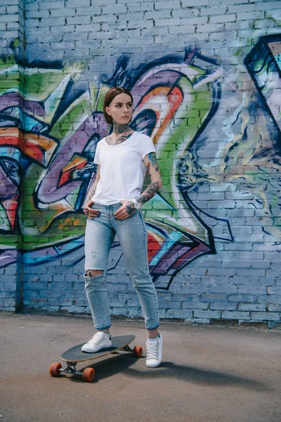 Молодая Татуированная Женщина Стоящая Скейтборде Возле Стены Красочными Граффити — стоковое фото