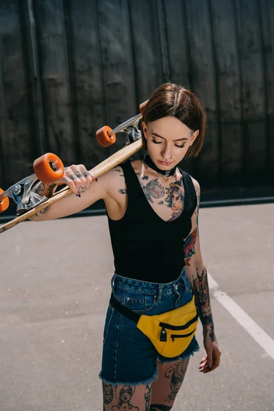 stylish tattooed girl with closed eyes holding skateboard over shoulder