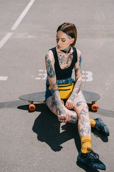 stylish young woman with tattoos sitting on skateboard at parking lot
