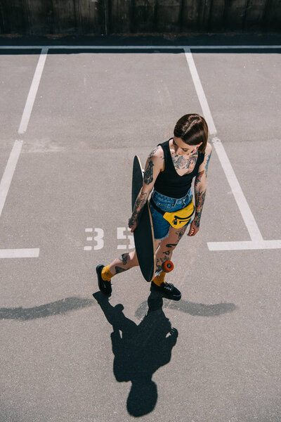 high angle view of stylish tattooed girl holding skateboard at parking lot 