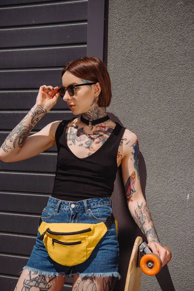 Молодая Стильная Женщина Татуировками Регулирующая Солнцезащитные Очки Стоящая Скейтбордом — Бесплатное стоковое фото