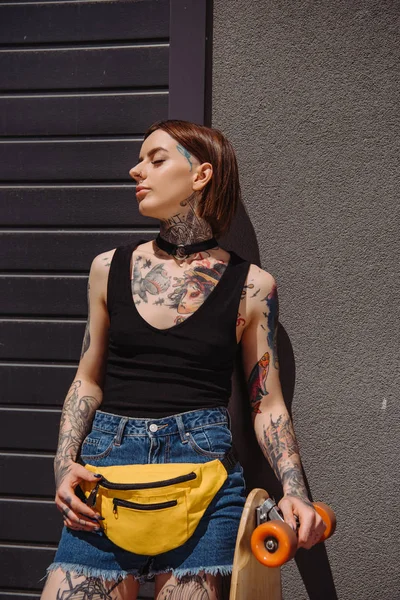 stylish tattooed woman with closed eyes standing with skateboard