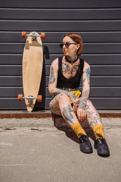 stylish woman with tattoos sitting near skateboard at street