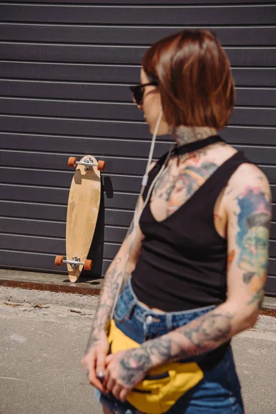 young tattooed woman listening music in earphones and looking at skateboard near wall