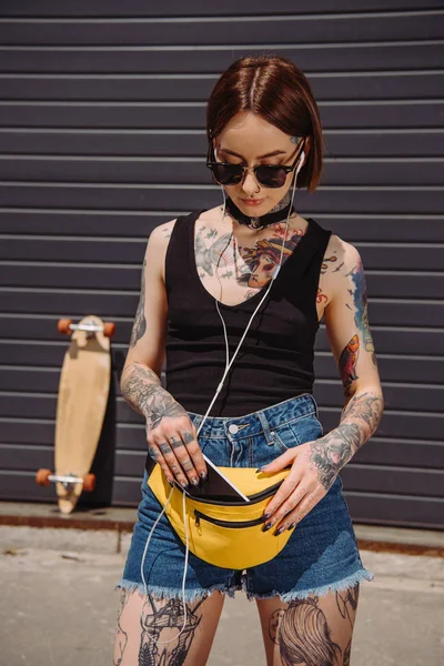 Stylish Tattooed Woman Listening Music Earphones Putting Smartphone Waist Bag — Stock Photo, Image