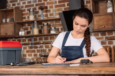 confident young repairwoman in overall writing in clipboard at kitchen clipart