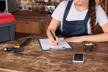 cropped shot of young repairwoman in overall writing in clipboard at kitchen clipart