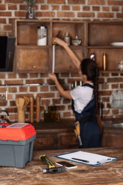 tools lying on kitchen table with blurred young repairwoman measuring cabinet on background clipart