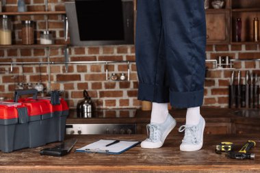 cropped shot of repairwoman in work pants and white shoes standing on toes on kitchen table clipart