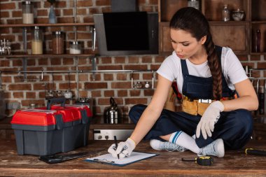 beautiful young repairwoman sitting on kitchen table with tools and writing in clipboard clipart