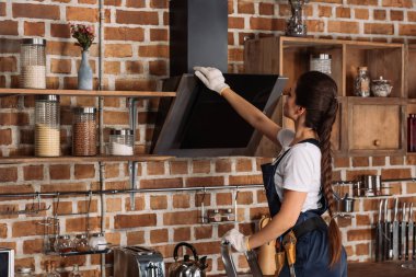 young repairwoman in overall touching stove hood at kitchen clipart