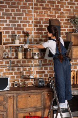 young repairwoman in overall measuring shelf at kitchen clipart