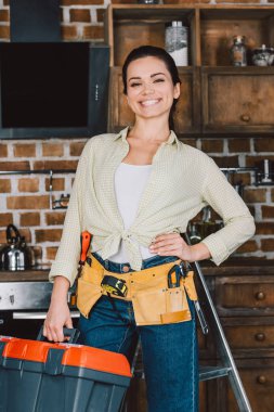 mutlu genç repairwoman toolbelt ve mutfak ayakta ve kameraya bakarak kutusu