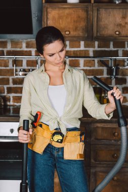 confused young repairwoman looking at vacuum cleaner at kitchen clipart