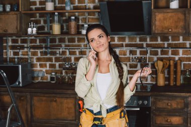 expressive young repairwoman talking by phone and looking up at kitchen clipart