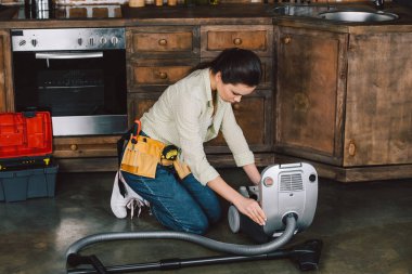 attractive young repairwoman fixing vacuum cleaner on floor of kitchen clipart