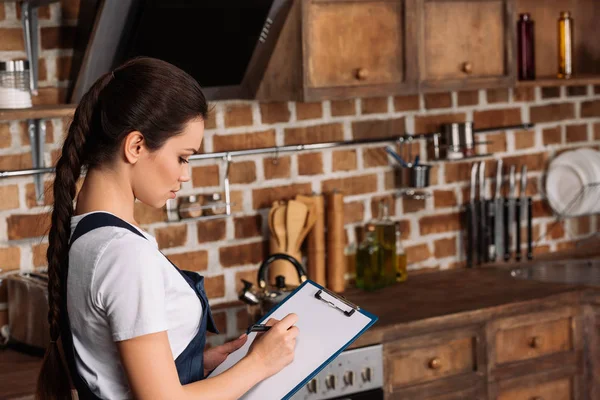 Selbstbewusste Junge Reparaturfrau Schreibt Klemmbrett Während Sie Der Küche Steht — Stockfoto