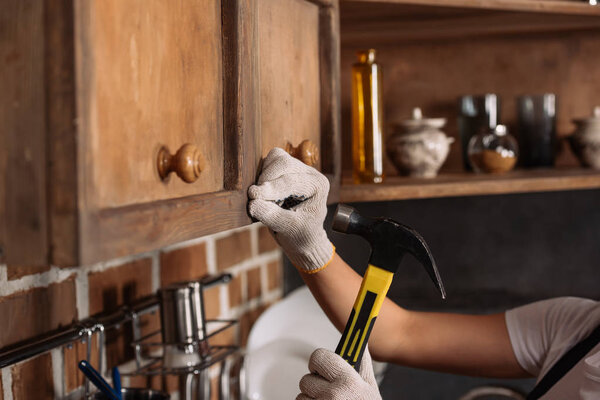 cropped shot of repairwoman in work gloves hammering nail into kitchen cabinet