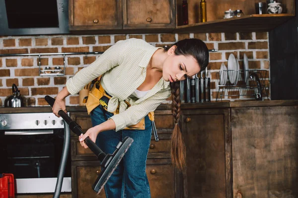 confused young woman with toolbelt looking at vacuum cleaner pipe at kitchen