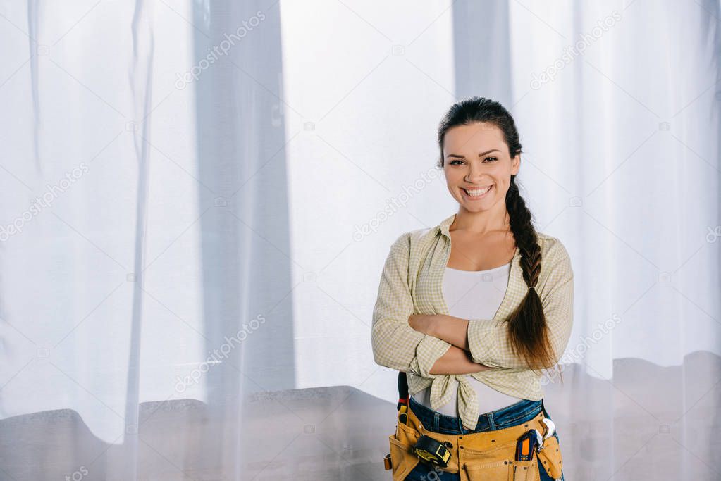 beautiful young repairwoman with toolbelt and crossed arms looking at camera