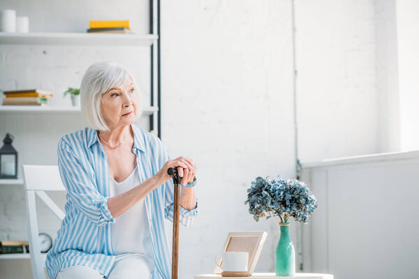 portrait of thoughtful senior woman with walking stick looking away at home