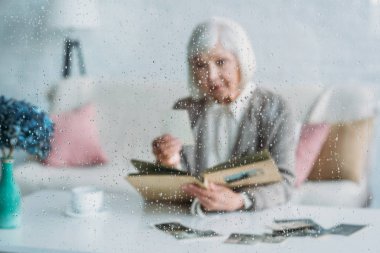 selective focus of senior woman looking at photographs in photo album at table with cup of coffee at home clipart