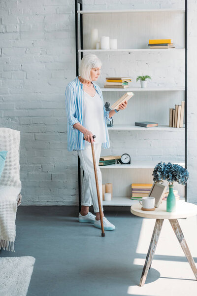 side view of grey hair woman with walking stick and book standing near bookshelf at home