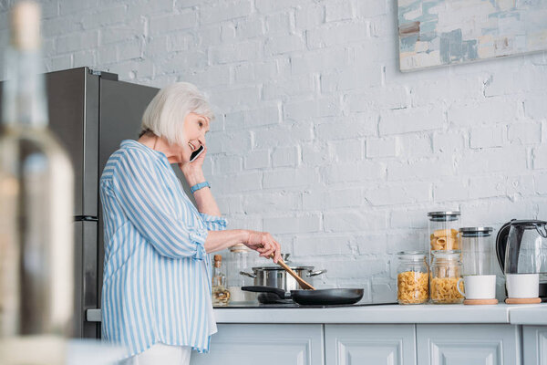 side view of smiling senior woman talking on smartphone while cooking dinner in kitchen