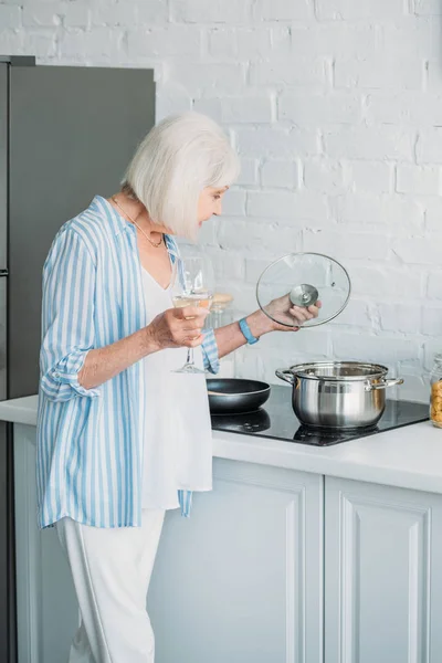 キッチンのストーブの上に鍋をチェック ワインのグラスとシニア女性の側面図  — 無料ストックフォト