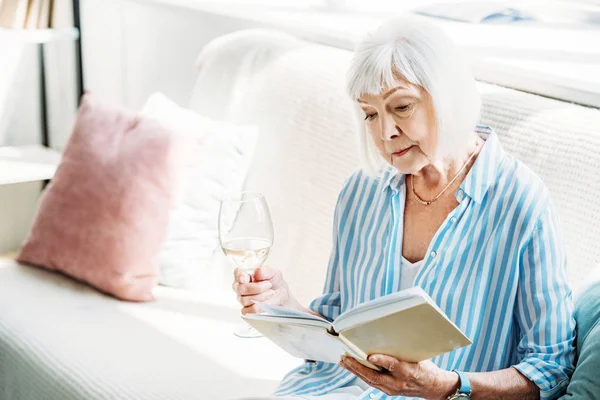 Сосредоточенная Пожилая Женщина Бокалом Вина Чтение Книги Диване Дома — стоковое фото