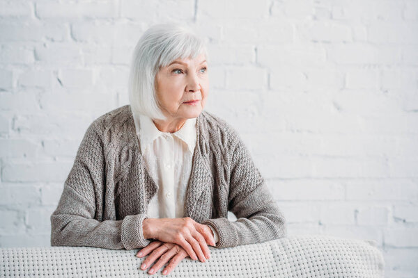 portrait of grey hair lady in knitted jacket leaning on couch and looking away at home