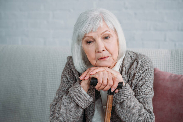 portrait of thoughtful grey hair lady with walking stick sitting on sofa at home