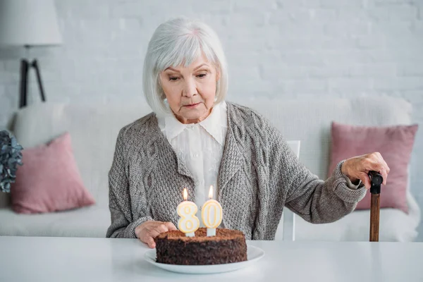 沉思的资深夫人坐在桌与生日蛋糕与蜡烛单独在家 — 图库照片