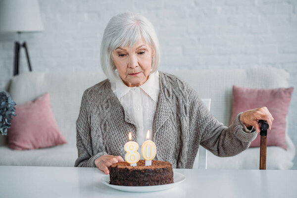 задумчивая старшая леди сидит за столом с тортом на день рождения со свечами в одиночестве дома
