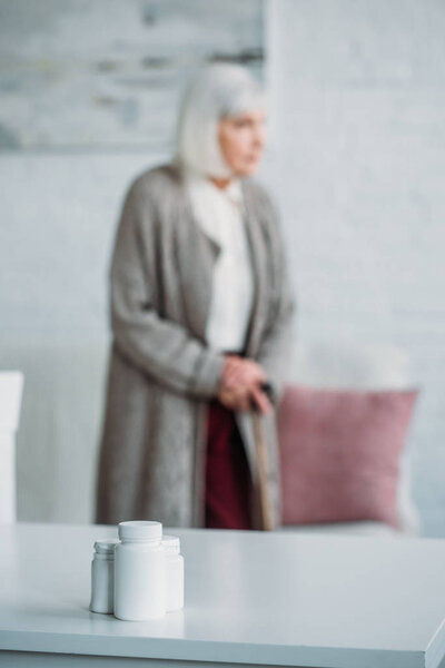 selective focus of senior woman with walking stick standing in room with medicines on tabletop at home