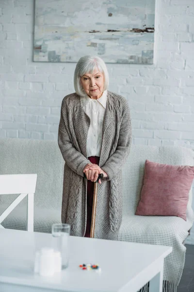 grey hair woman with walking stick standing in room with medicines on tabletop at home