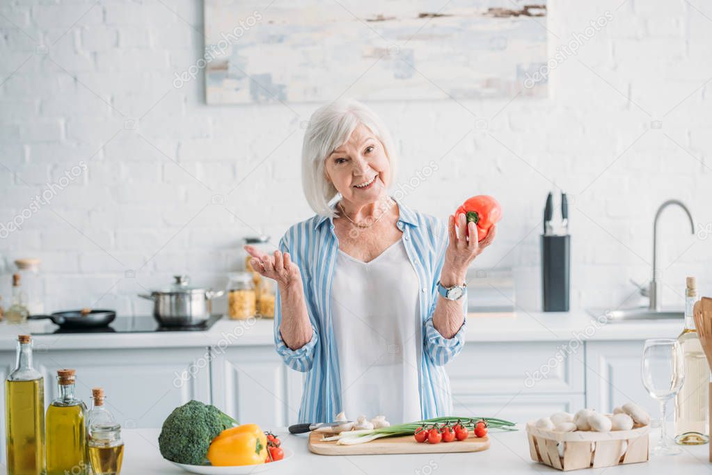 portrait of smiling grey hair lady with bell pepper in hand standing at counter in kitchen