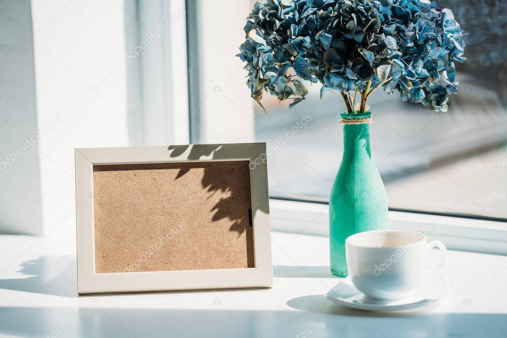 close up view of empty photo frame, cup of coffee and bouquet of hortensia flowers in vase on windowsill